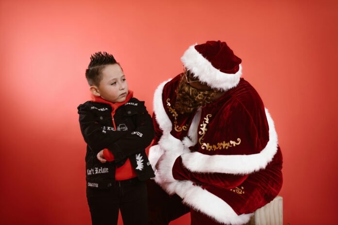 A young child interacts with a Santa Claus in festive attire against a red backdrop.